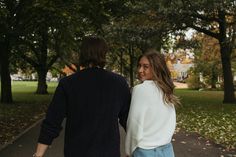 a man and woman holding hands walking down a path in the park with leaves on the ground