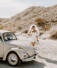 a woman in a wedding dress standing next to a vw bug on the beach