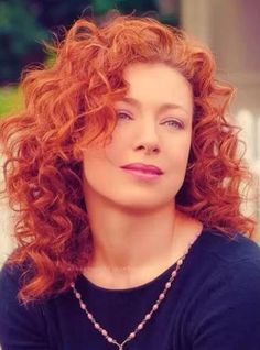 a woman with red curly hair wearing a black shirt and necklace looking up at the camera