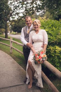 a bride and groom pose for a photo on the side of a wooden fence with greenery in the background