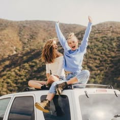 two women sitting on the roof of a car with their arms up in the air