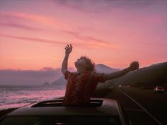 a person sitting on top of a car with their arms up in the air as the sun sets