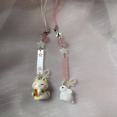 two key chains with charms attached to them on a white cloth covered surface, one has a cat and the other is a dog