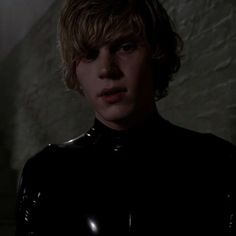 a young man in a black leather outfit looks at the camera while standing in a dimly lit room