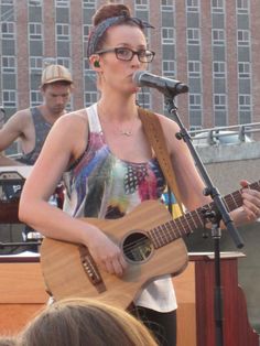 a woman with glasses playing an acoustic guitar in front of a microphone and some people