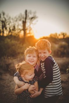 three young children are hugging each other in the desert at sunset, with trees and bushes in the background