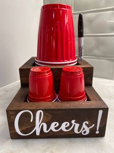 a wooden tray with two red cups and a sign that says cheers on it, sitting on a counter