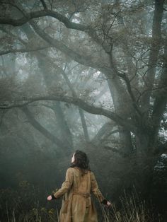 a woman standing in the middle of a field with trees behind her and foggy skies overhead