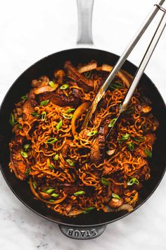 a wok filled with noodles and meat on top of a white counter next to two tongs