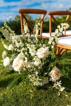 white flowers and greenery sit on the grass near two wooden chairs in the background