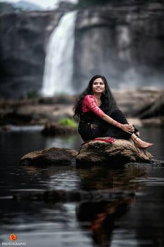 a woman sitting on top of a rock next to a waterfall in the middle of a body of water