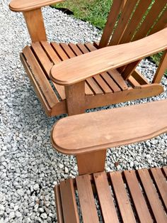 two wooden chairs sitting next to each other on top of gravel covered ground with grass in the background