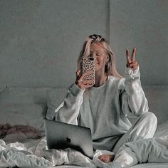 a woman sitting on a bed holding a cell phone and looking at her laptop computer