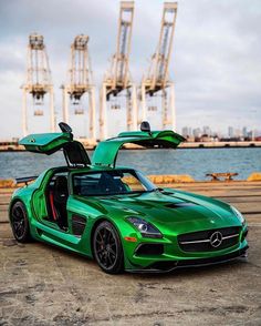 a green sports car with its doors open sitting in front of the water and cranes