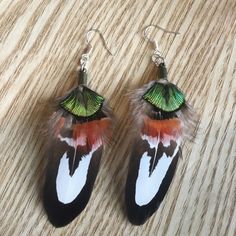 a pair of earrings with feathers and green leaves on them sitting on a wooden surface