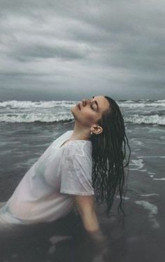 a woman laying on the beach with her head in the water and looking up at an overcast sky