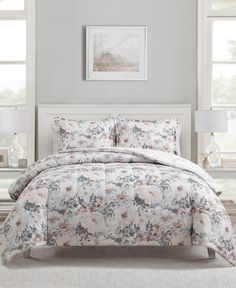 a white bed with floral comforter and pillows in front of two large window frames