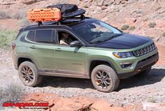 a green jeep with an orange toolbox on top is parked in the dirt near some rocks