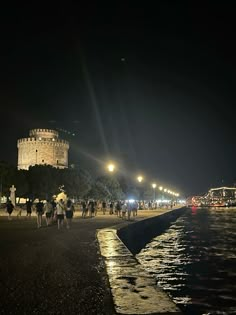 people are walking along the water at night near a white castle with its lights on