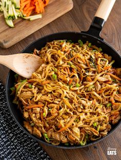 a pan filled with noodles and vegetables on top of a wooden table next to a cutting board