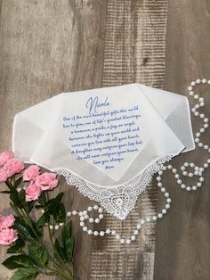 a white handkerchief with blue writing on it next to pink flowers and beads, along with a string of pearls