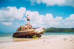 a man standing on top of an old tank in the sand at the beach with blue sky and clouds behind him