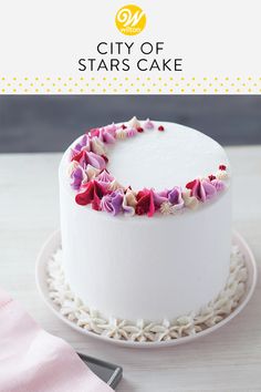 a white cake with pink and purple flowers on the top is sitting on a plate