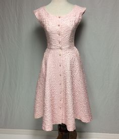 This very unusual true vintage 1950's pink quilted cotton dress has a fitted bodice, scooped neckline, cap sleeves and a flaired skirt. The fabric has a sheen to it and is backed with gauze. Two darts in the front and two princess seams in the back give the bodice a very fitted look. The front closure is created by 11 pink and rhinestone buttons and button holes, and a waistline snap. The flaired skirt has two side pockets. Some stitching is slightly loose on the left top shoulder. I have found no stains or discolorations.  Label reads "Alix of Miami." Measurements:      Sleeve cap to sleeve cap: 18 1/4"      Bust: 35"      Waist: 26 1/4"      Hips: free      Front shoulder to waist: 16"      Front shoulder to hem: 42 1/2"      Waist to hem: 26 1/4"      Skirt circumference: 120"      Fron Fitted Vintage Dress With Buttons, Spring Vintage Fitted Dress With Button Closure, Fitted Vintage Dress With Button Closure For Spring, Pink Fitted A-line Vintage Dress, 1950s Style Fitted Dress With Buttons, Fitted Vintage Dress With Buttons For Garden Party, Pink Party Dress With Button Closure, Fitted Button Dress For Garden Party, Fitted Tea-length Vintage Cotton Dress