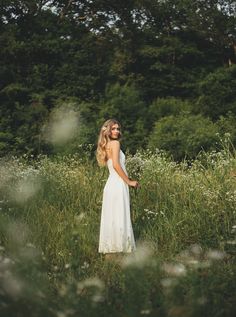 a woman in a white dress is standing in tall grass with trees in the background