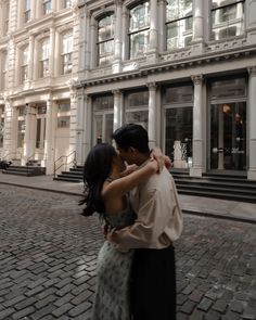 a man and woman kissing on the street in front of a tall building with many windows