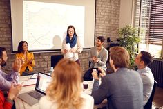 a group of people sitting around a table in front of a projector screen with a woman giving a presentation