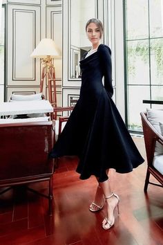 a woman in a black dress standing next to a chair and lamp on a hard wood floor