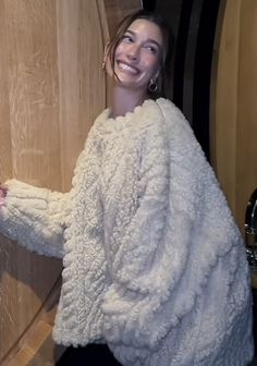 a woman in a fuzzy white coat smiles as she opens the door to her home