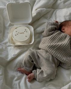 a baby laying on top of a white blanket next to a cake in a box