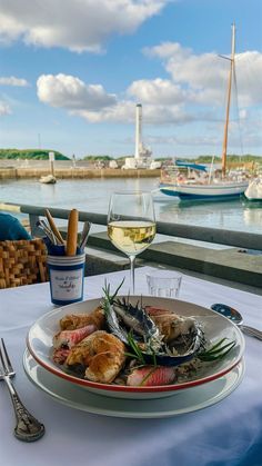 a plate of food on a table next to a glass of wine and some boats in the water