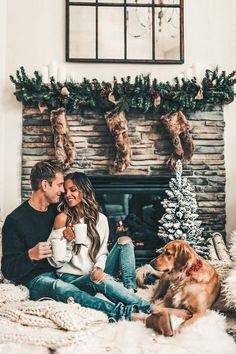 a man and woman sitting next to a dog in front of a fireplace with christmas decorations