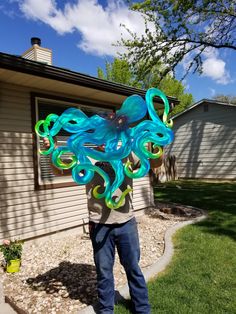 a man standing in front of a house holding up an octopus kite with blue and green swirls on it