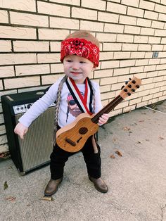 a small child wearing a red bandana and holding a ukulele guitar in front of a brick wall