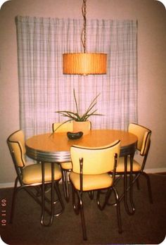 a dining room table with four chairs and a plant on the center piece, in front of a curtained window