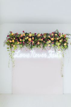 a neon sign that says love never stops with flowers hanging from the wall behind it