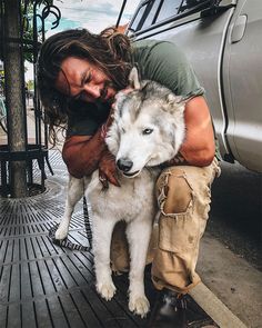 a man is hugging his wolf dog on the ground next to a car and fence