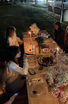 a group of people sitting around a table with food and candles on it at night