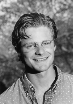 a black and white photo of a man with glasses smiling at the camera in front of a tree