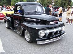 an old black pickup truck parked in a parking lot next to other cars and people