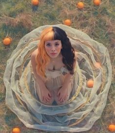 a woman with long hair sitting on the ground surrounded by oranges and mesh netting