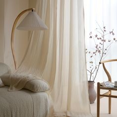 a white bed sitting next to a window covered in sheer curtains and a vase filled with flowers