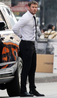 a man standing next to a parked car wearing a white shirt and black tie with his hands in his pockets