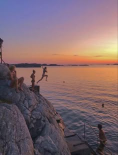 some kids are playing on the rocks by the water at sunset or sunrise, while others swim in the water