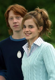 two young people standing next to each other in front of green trees and bushes, one wearing a blue sweater