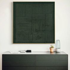 a black dresser with a plant on top and a green wall hanging above it in a white room